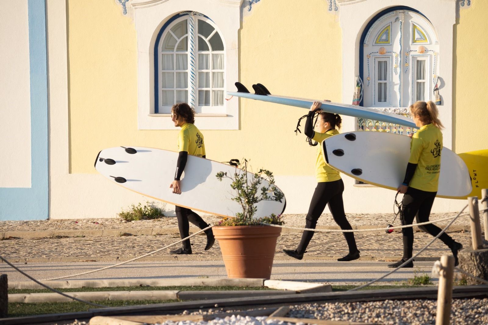 Three people carrying soft surfboards beside a yellow tiled house in Ericeira